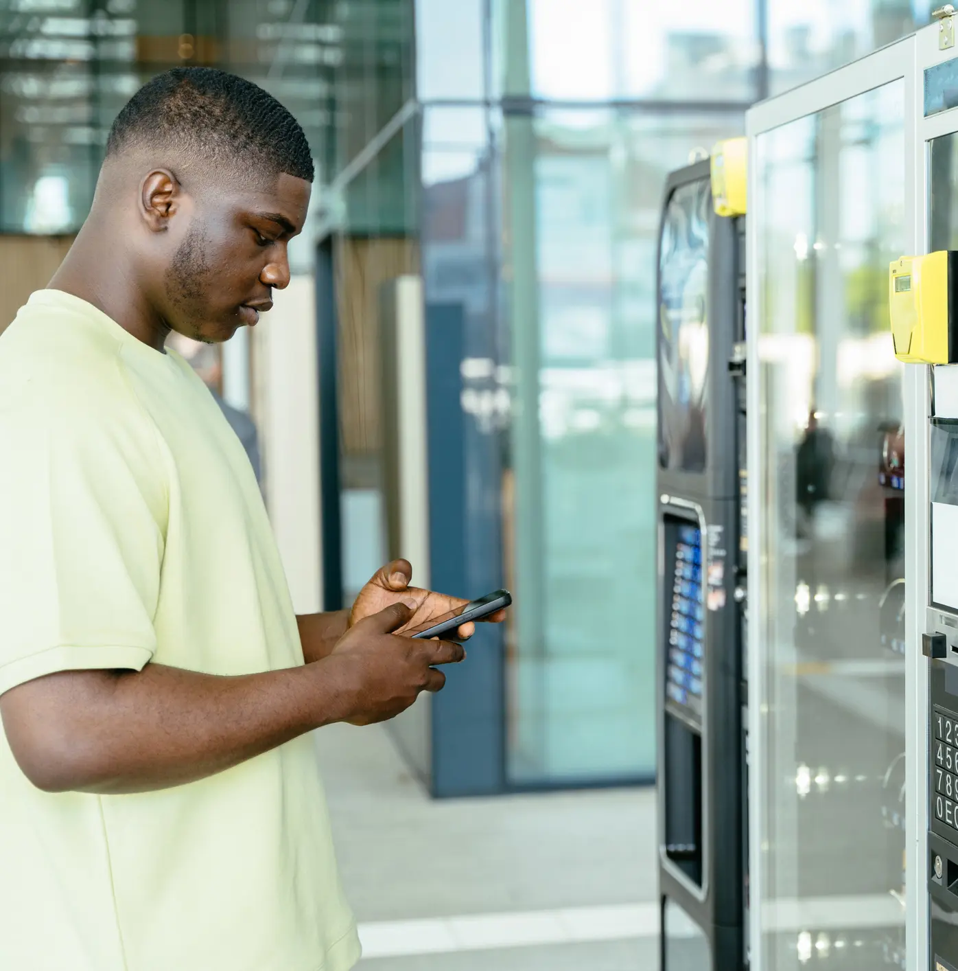 Young man using phone next to vending machines in a modern building.